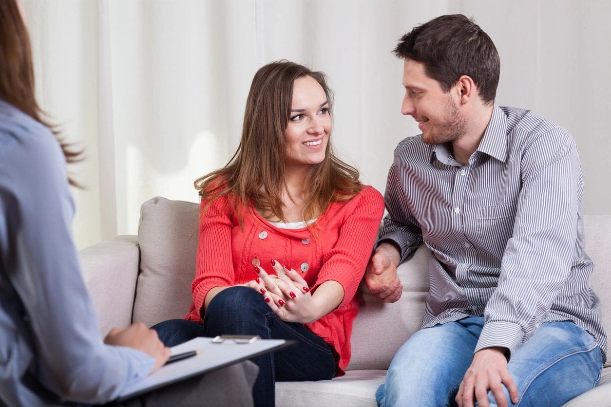 A man and woman sitting on the couch talking to another man.